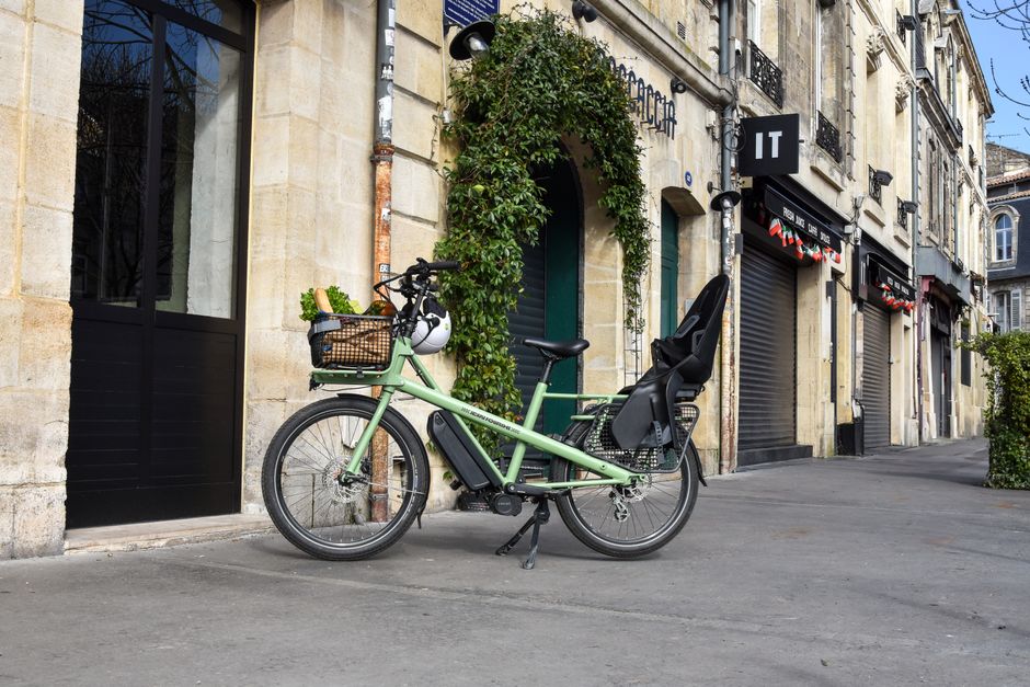A green cargo e-bike with groceries parked outside a French building on a sunny day.