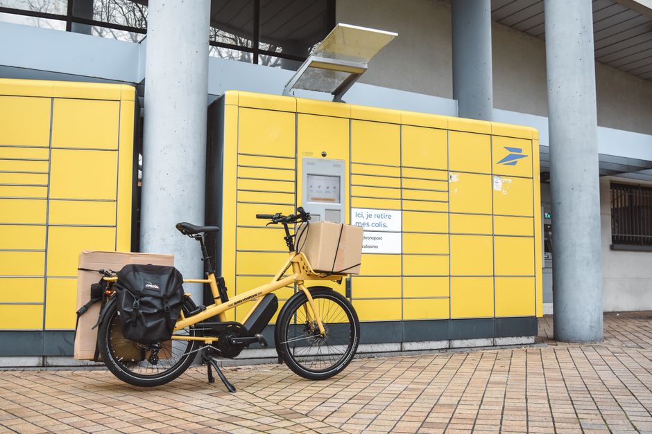 A yellow cargo e-bike loaded with parcels parked outside a package pickup station in France.